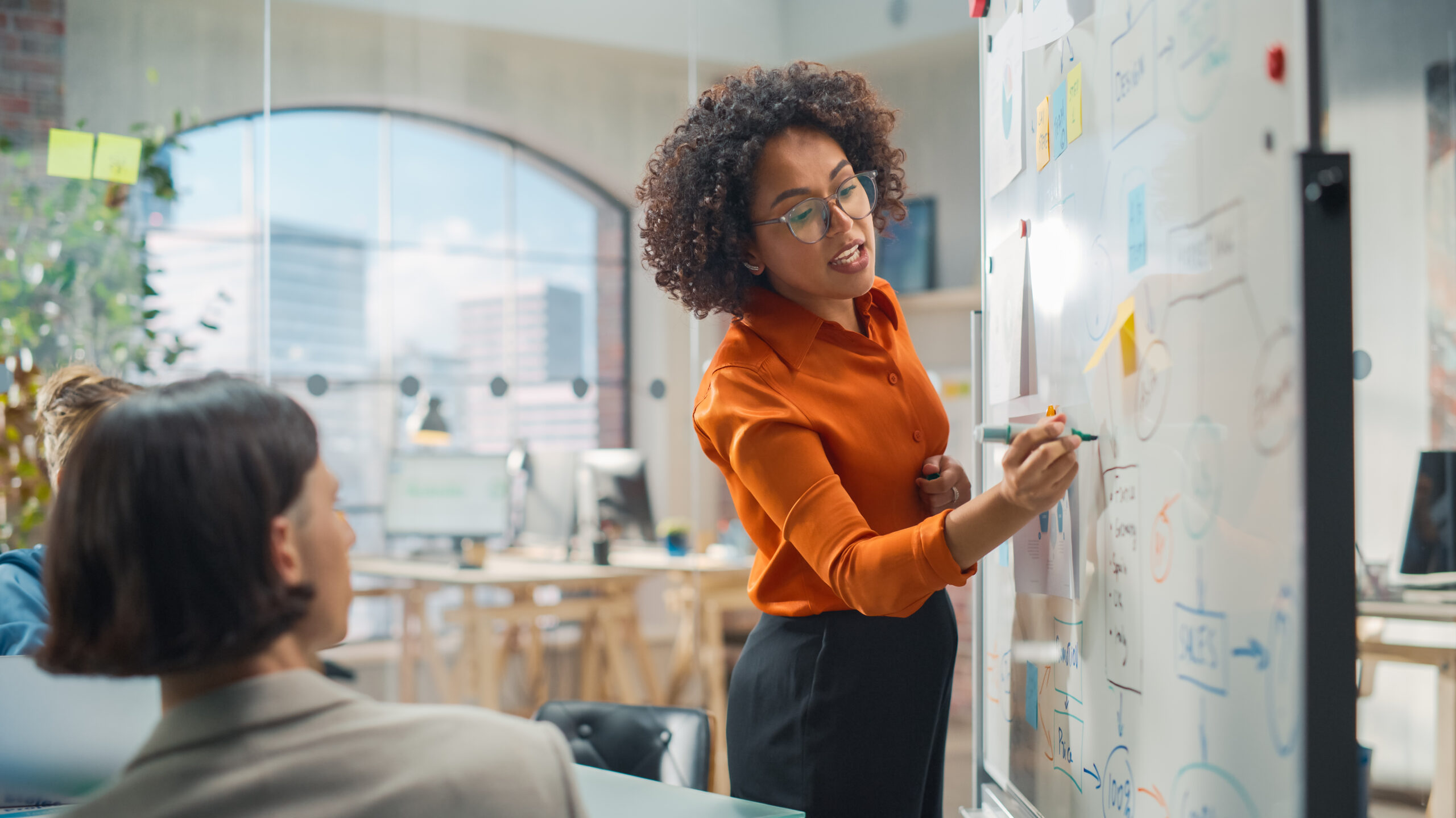 A woman writing on a whiteboard while standing in front of her coworkers. She is wearing professional, tidy clothes.