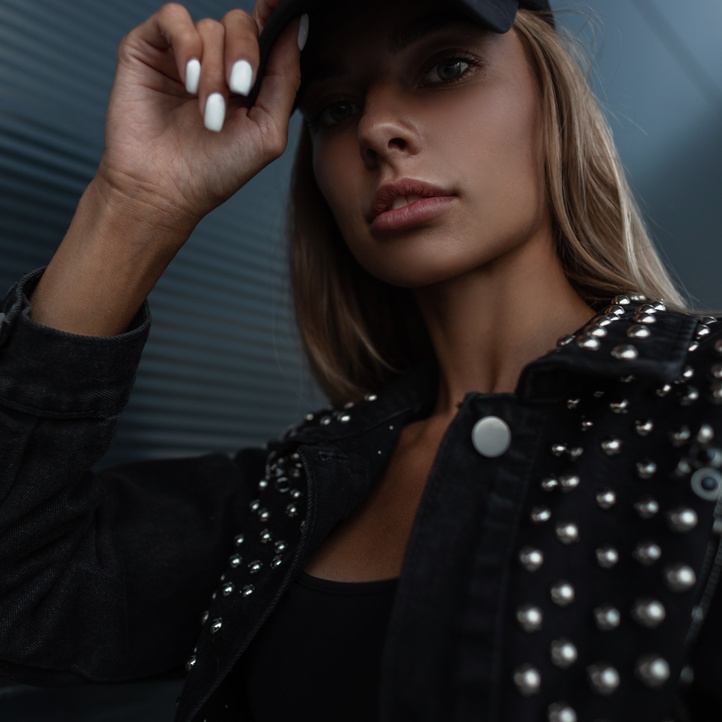 A woman models a black baseball cap, holding her hand to the brim, while dressed in all black against a gray background.
