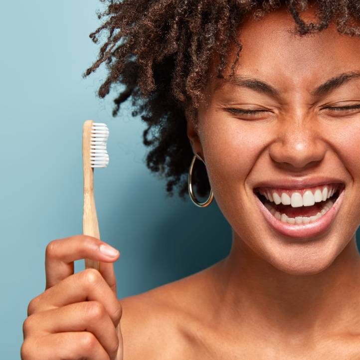 A woman holds a wooden toothbrush beside her head and smiles, her eyes closed and her teeth in full view.