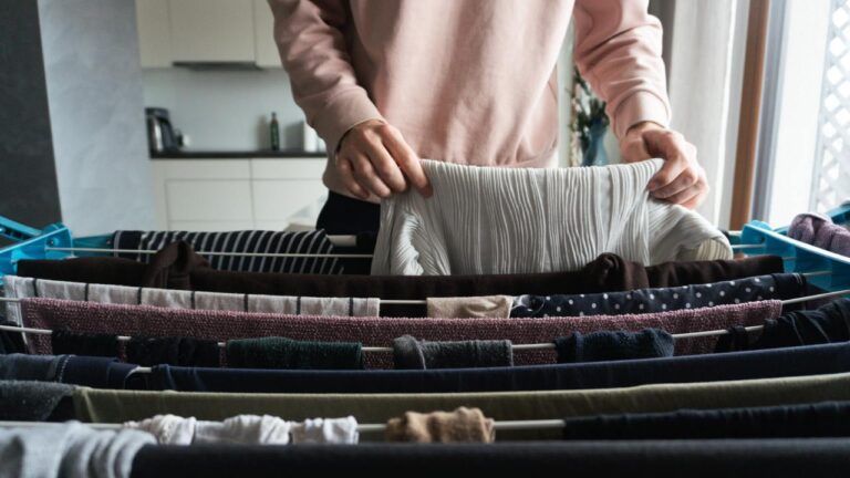 A person places a white sweater on a wire drying rack. Several other garments are drying on the rungs.