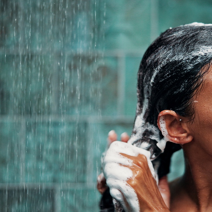 A close-up of a woman in a shower with green-blue-tiled walls, washing her black hair with foaming soap.