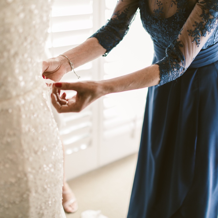 A mother of the bride in a floor-length blue gown with lace sleeves helps secure the buttons on her daughter's wedding dress.