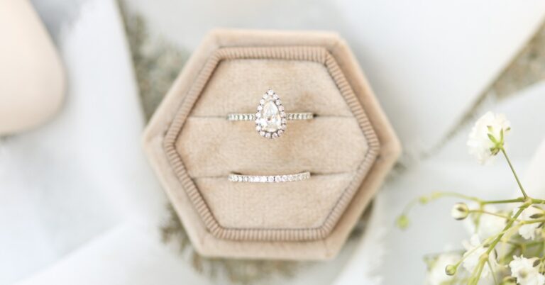 Pear-shaped diamond engagement ring and matching wedding band in a beige velvet box beside delicate white flowers.