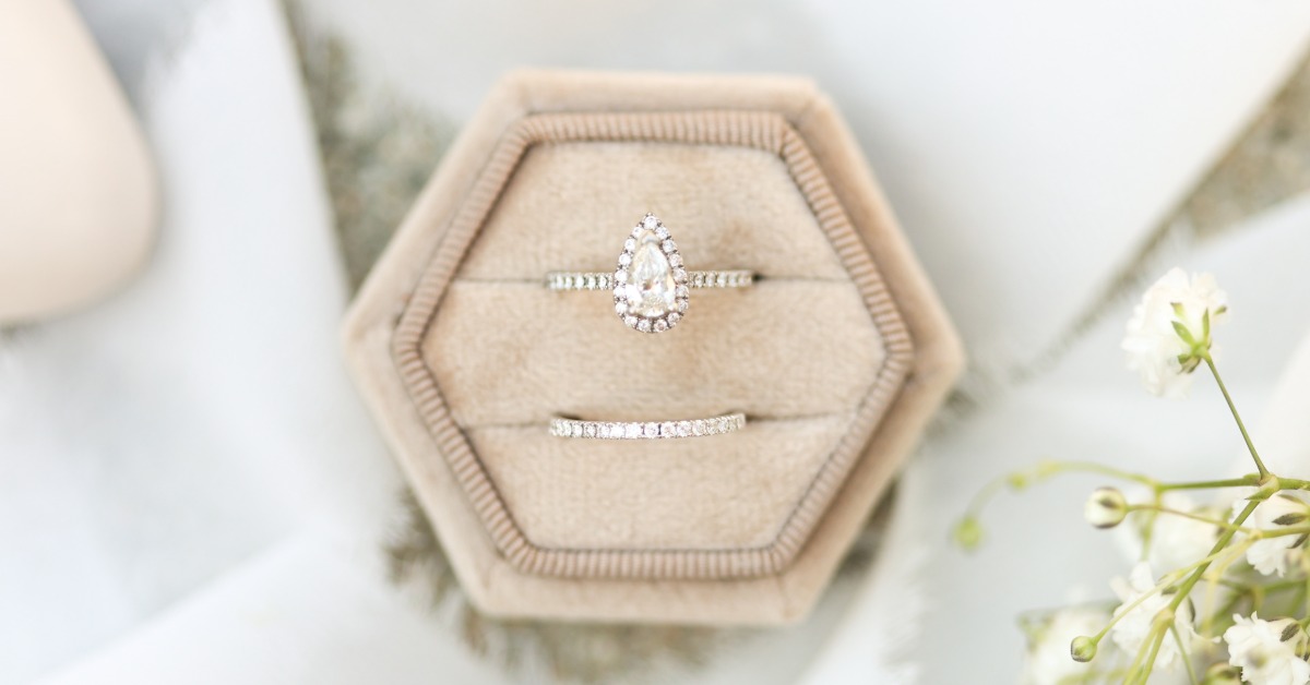 Pear-shaped diamond engagement ring and matching wedding band in a beige velvet box beside delicate white flowers.