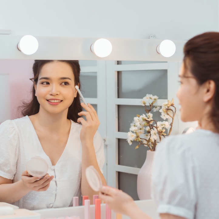 A woman applying makeup with a brush while facing a lit vanity mirror beside flowers and cosmetic items.