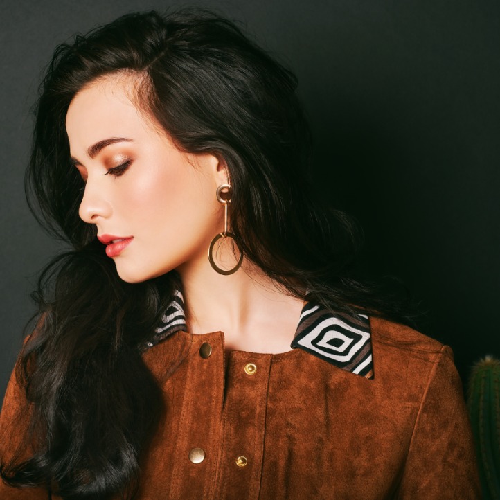 A woman with long, dark hair wears a brown suede jacket and hoop earrings while posing beside a tall cactus.