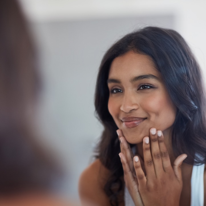 A smiling woman gently touches her face while looking in a mirror, showing smooth, healthy-looking skin.