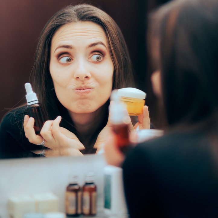 A brunette woman in the bathroom mirror criticizes her skin's texture. She holds a few products up to her face.