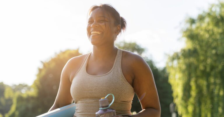 Smiling woman in workout clothes carries a rolled yoga mat and water bottle while standing outdoors in sunlight.