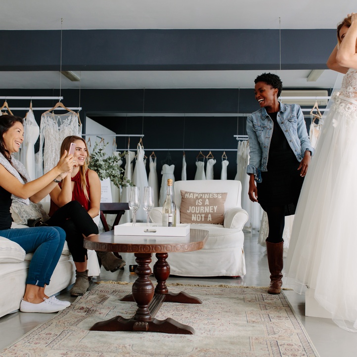 Bride wearing a white lace wedding gown and veil shows her dress to smiling loved ones inside a bridal boutique.