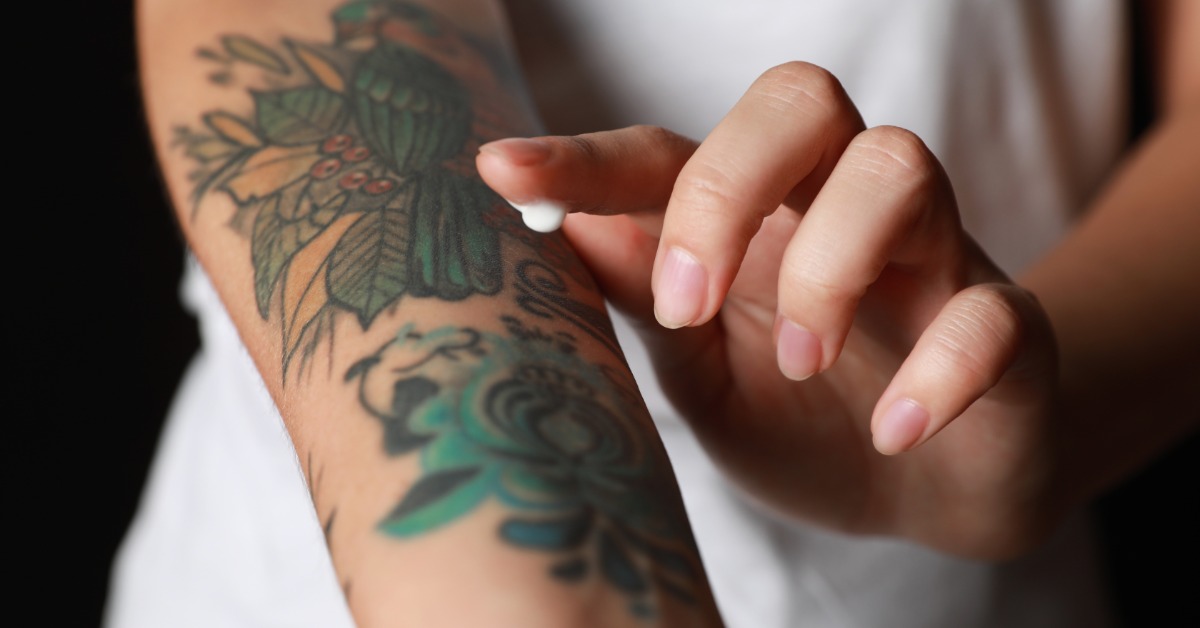 A close-up of the hand of a woman with a little drop of moisturizer on her finger, applying it over an arm tattoo.